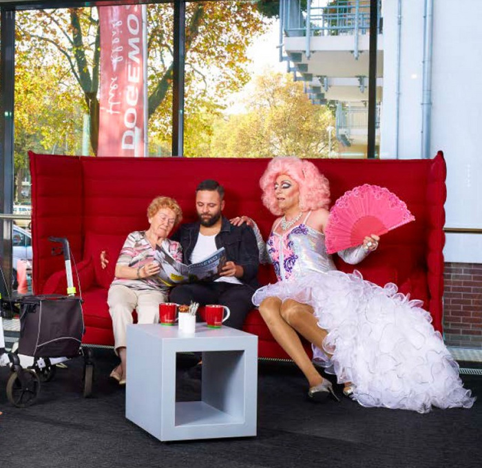 Eine Seniorin, ein Mann und eine Dragqueen sitzen gemeinsam auf einer roten Couch und blättern in einer Zeitung.