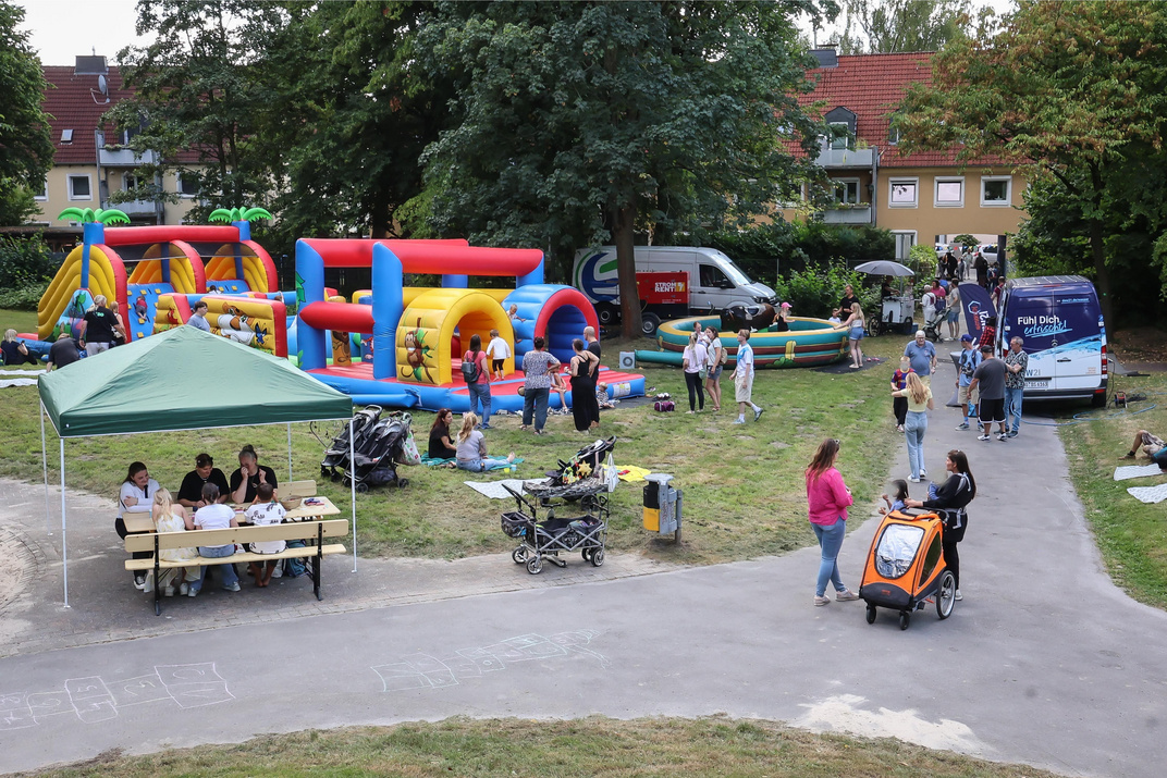 Auf einem Innenhof stehen auf der Grünfläche eine riesige Hüpfburg, ein Gerät zum Rodeoreiten und viele kleine und große Menschen sitzen auf Decken oder gehen über das Gelände.