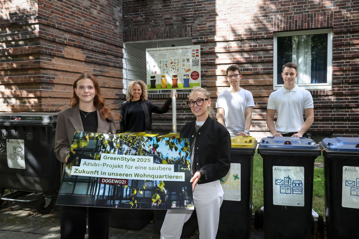 Fünf junge Menschen stehen vor einem Mehrfamilienhaus an einem Müllstandplatz, dahinter eine Stelle mit einem Schild mit Piktogrammen zur Mülltrennung.