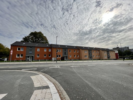 Blick von der gegenüberliegenden Straßenseite auf die vier energetisch modernisierten Mehrfamilienhäuser Am Hombruchsfeld 49-55, die unter einem tupfig bewölkten Himmel mit Sonne in frischen, rot-orangenen Farben strahlen