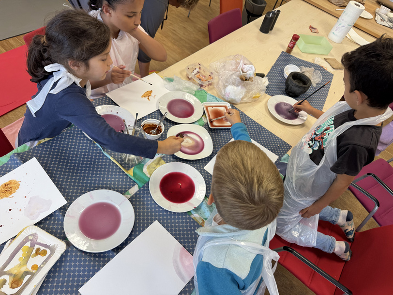Kinder sitzen an einem Tisch mit Schälchen voller Pflanzenfarben und malen.