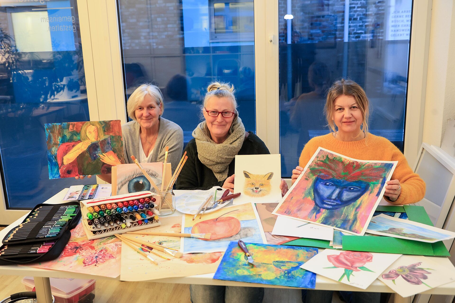 Drei Frauen zeigen bunte Zeichnungen und gemalte Bilder auf einem Tisch
