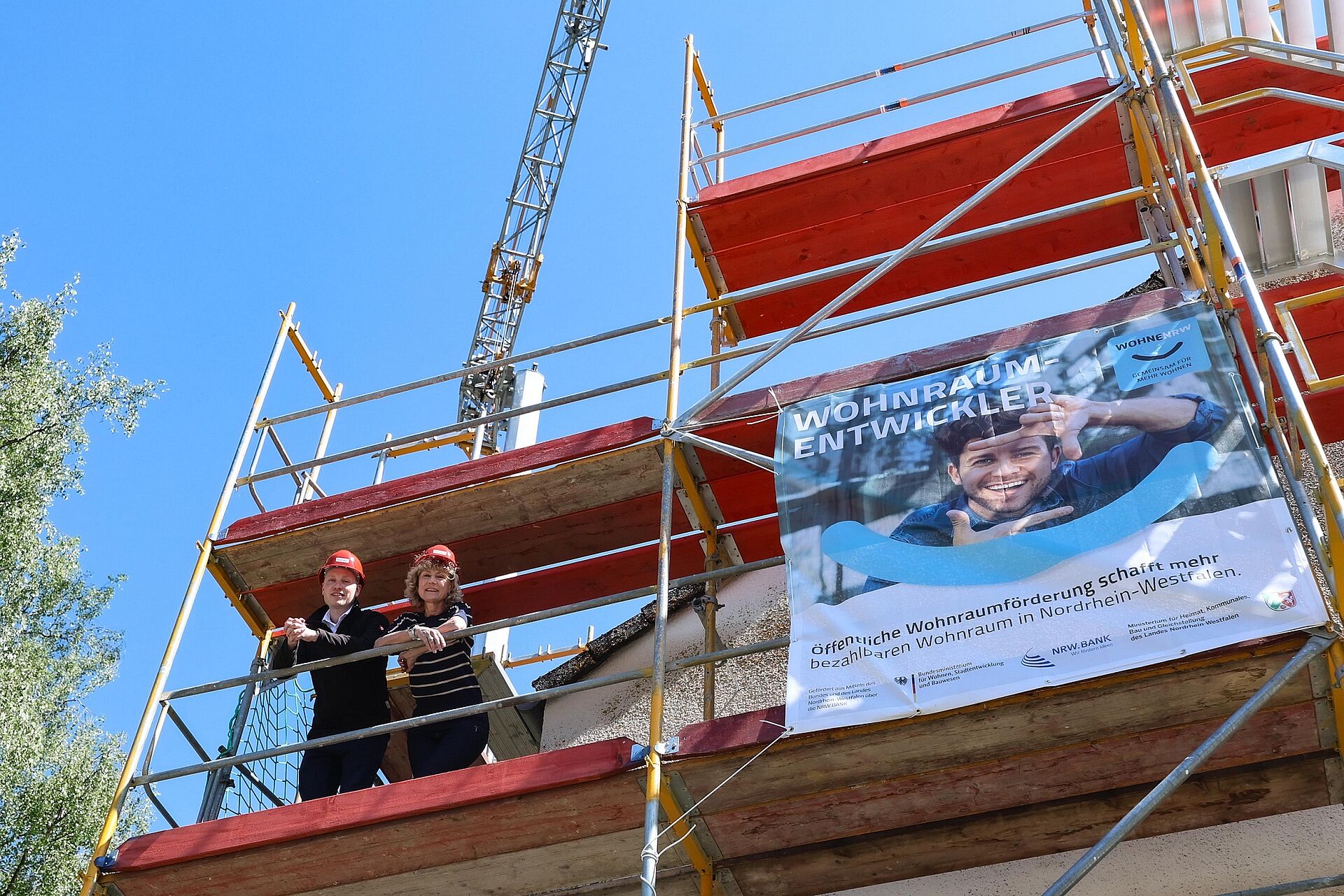Zwei Projektbeteiligte stehen auf dem Baugerüst eines eingerüsteten Mehrfamilienhauses, im Hintergrund strahlend baluer Himmel und am Gerüst hängt ein Banner mit den Förderern des Projektes.