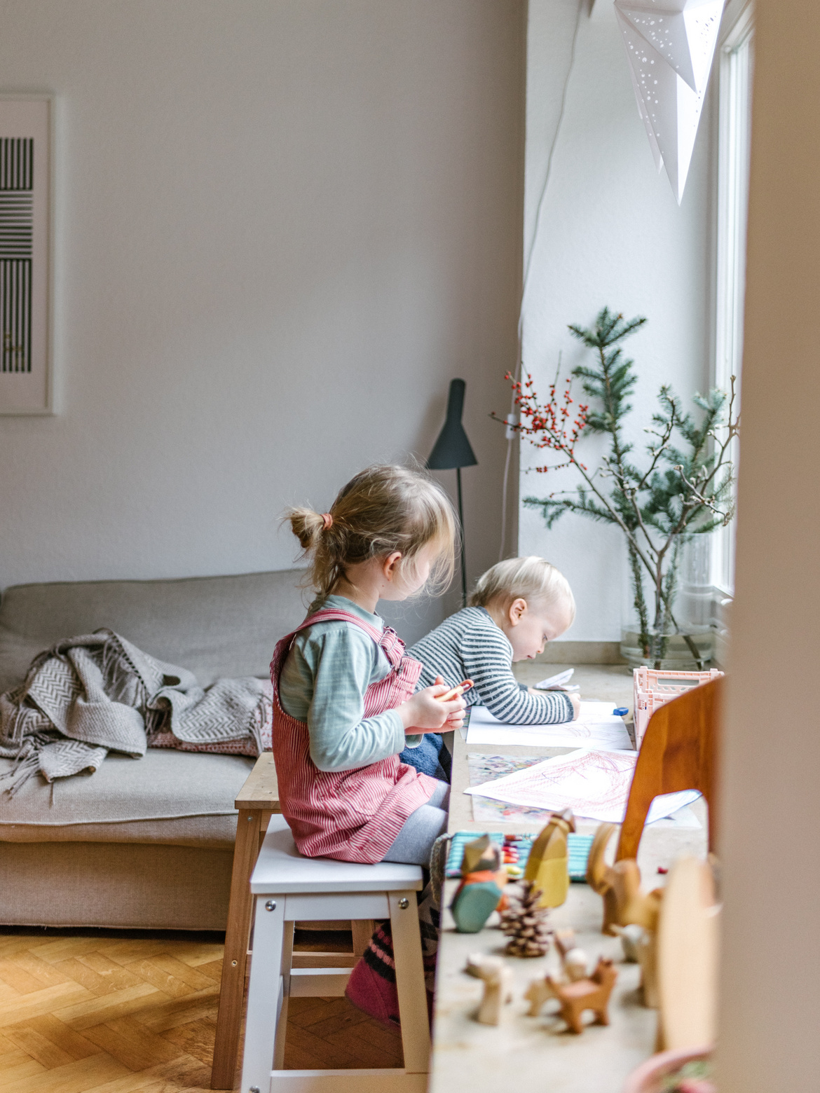 Zwei Kinder sitzen auf Holz-Hockern an einer breiten Fensterbank und malen.