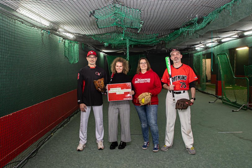 Zwei Frauen und zwei Männer stehen in einer Baseball-Trainingshalle mit Fangnetzen und halten einen symbolischen Spendenscheck.