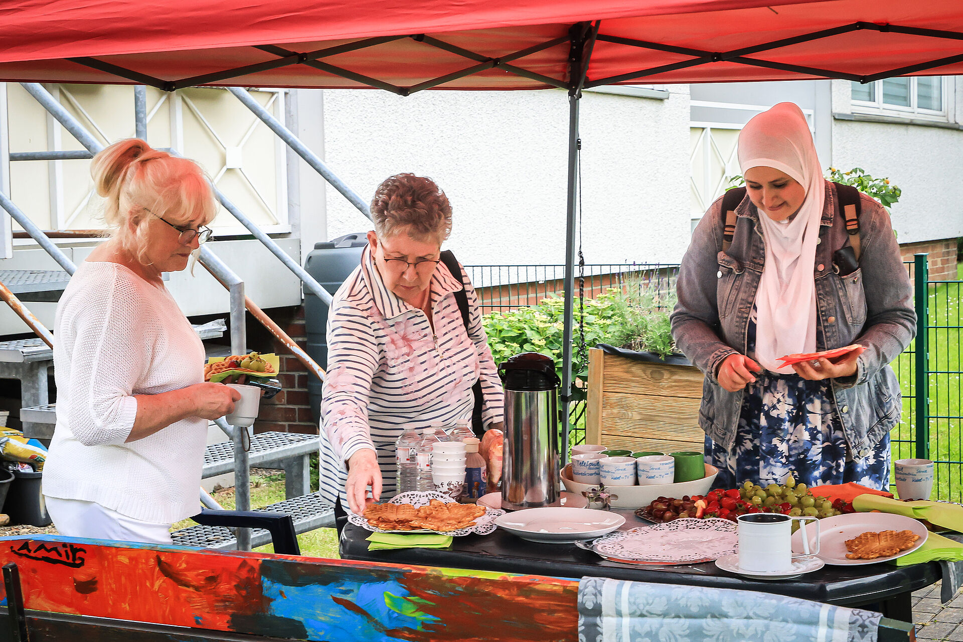Drei Frauen stehen an einem Tisch mit frischen Waffeln und Obst im Garten der Nachbarschaftsagentur.