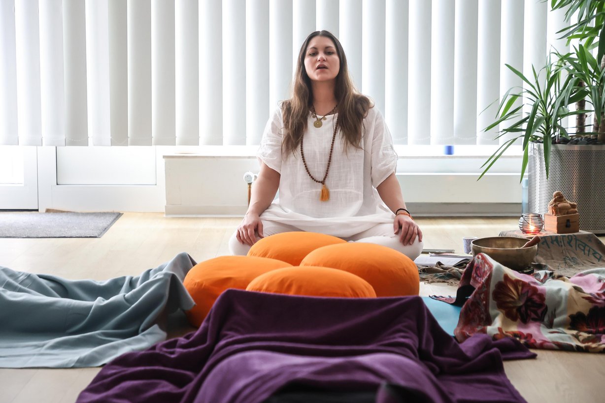 Eine Yogalehrerein sitzt auf einem Meditationskissen und führt Schülerinnen durch eine Yogaübung