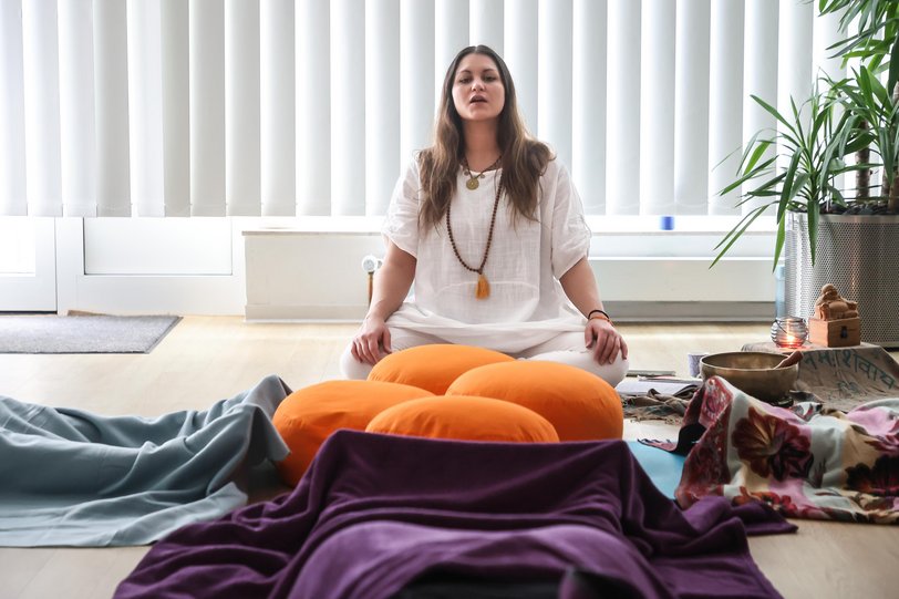 Eine Yogalehrerein sitzt auf einem Meditationskissen und führt Schülerinnen durch eine Yogaübung