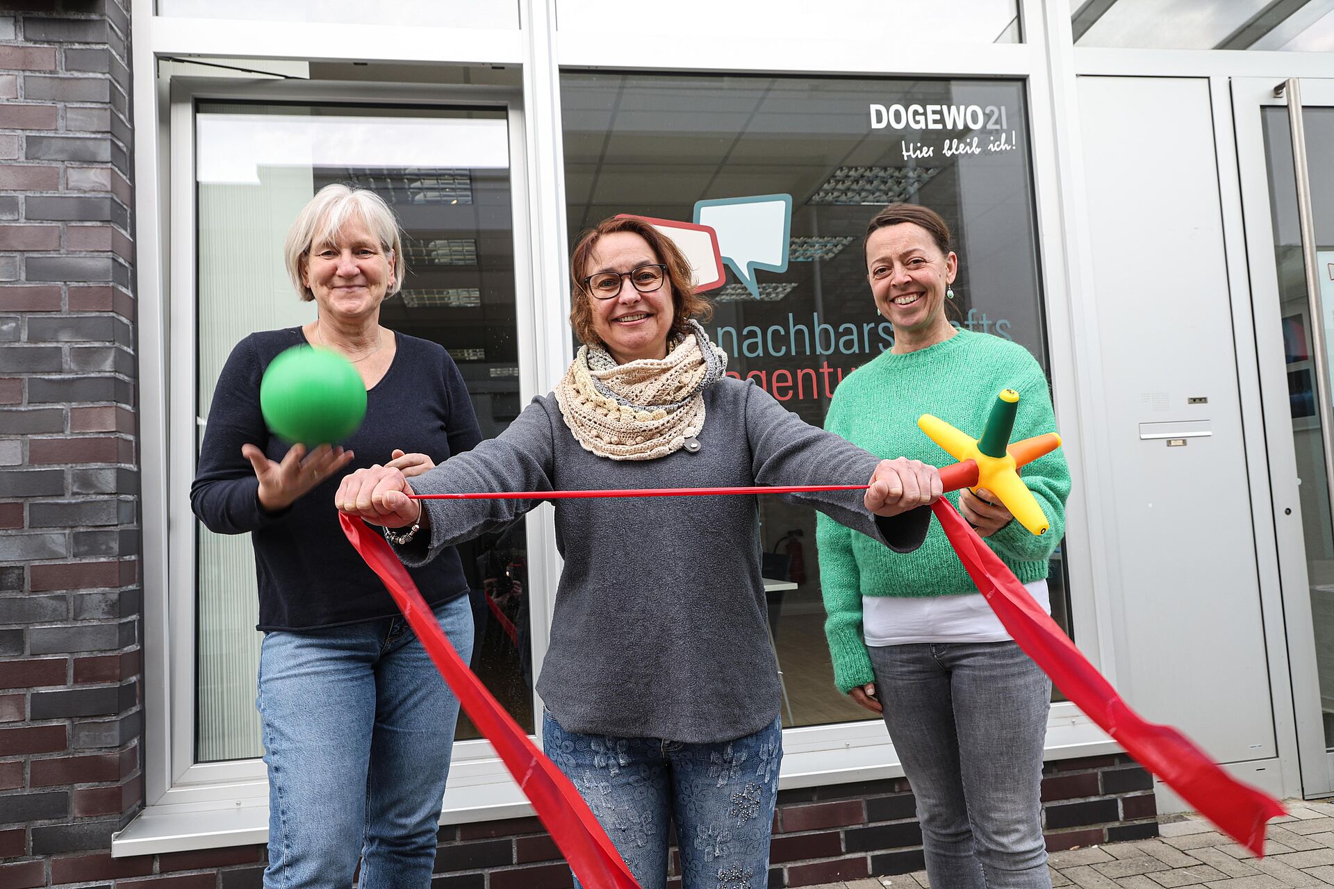 Drei Frauen stehen mit Terraband und Ball in den Händen vor der Nachbarschaftsagentur und schauen in Richtung Kamera