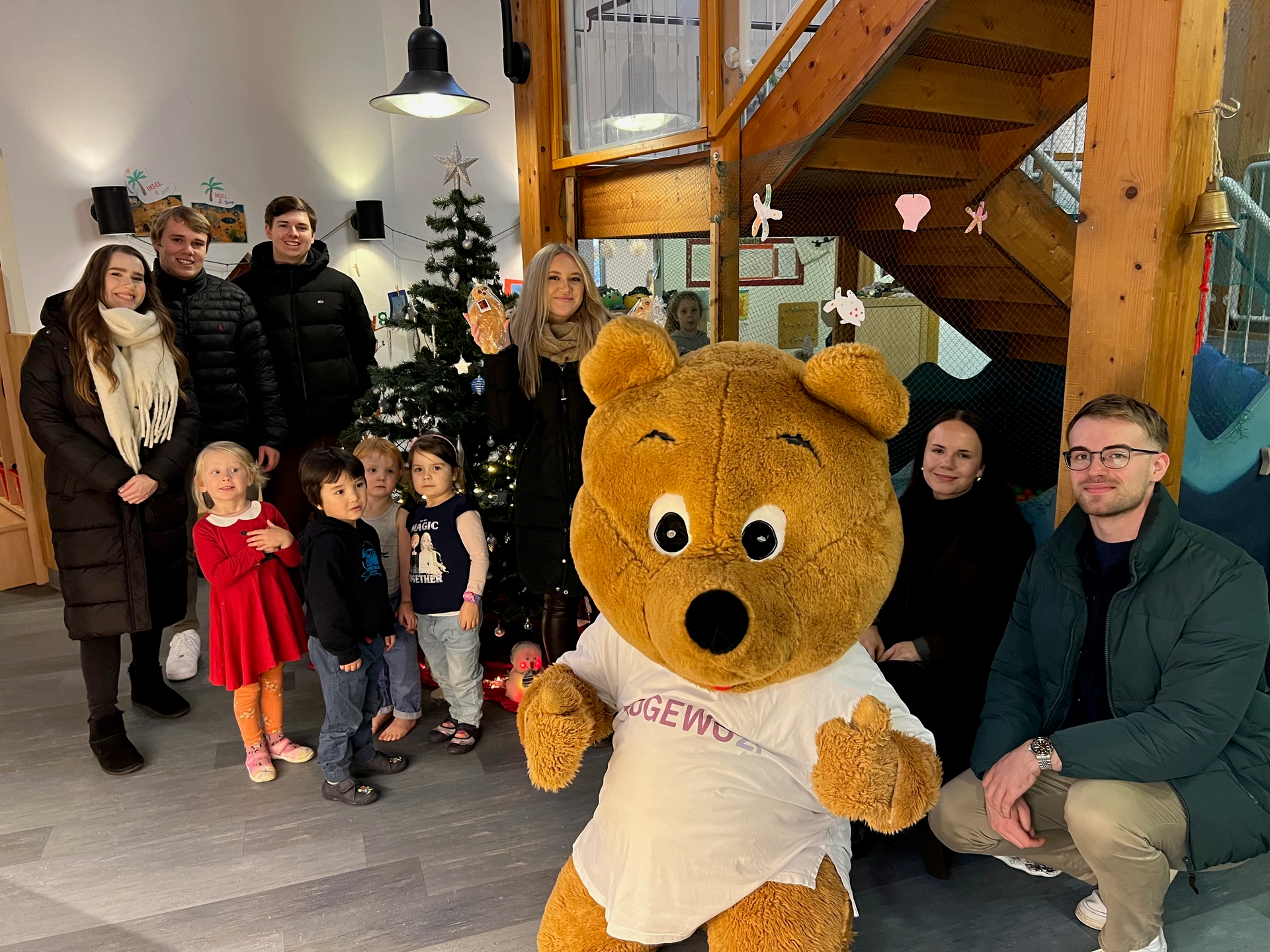Mehrere Kinder und junge Erwachsene lächeln gemeinsam mit ein lebensgroßes Bärenmaskottchen in einer Kindertageseinrichtung in Richtung des Fotografen