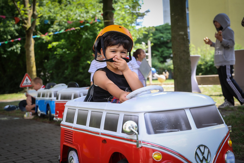 Ein Junge mit Helm fährt in einem Elektrokinderauto in einem grünen Innenhof und im Hintergrund stehen und spielen weitere Kinder.