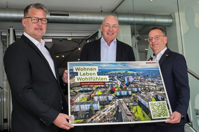 Drei Männer präsentieren im Eingangsbereich des DOGEWO21-Kundenzentrums ein großformatiges Titelbild des Geschäftsberichtes 2024.