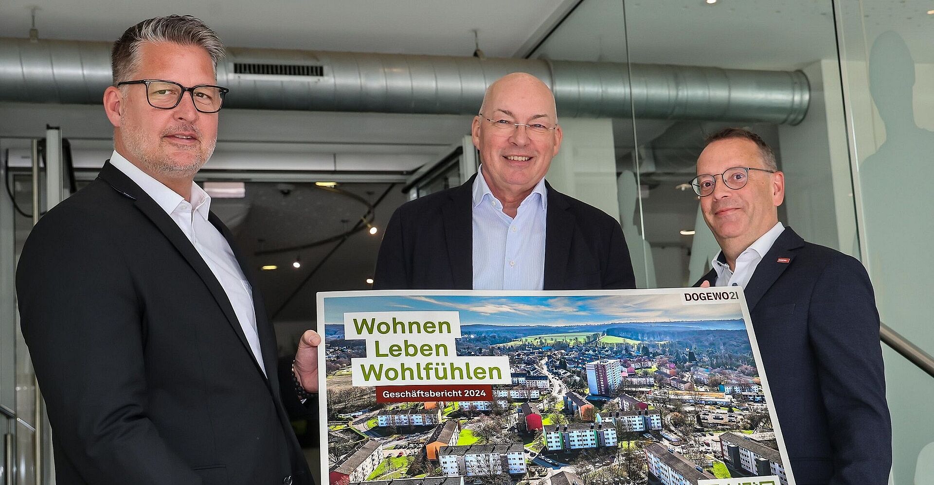 Drei Männer präsentieren im Eingangsbereich des DOGEWO21-Kundenzentrums ein großformatiges Titelbild des Geschäftsberichtes 2024.