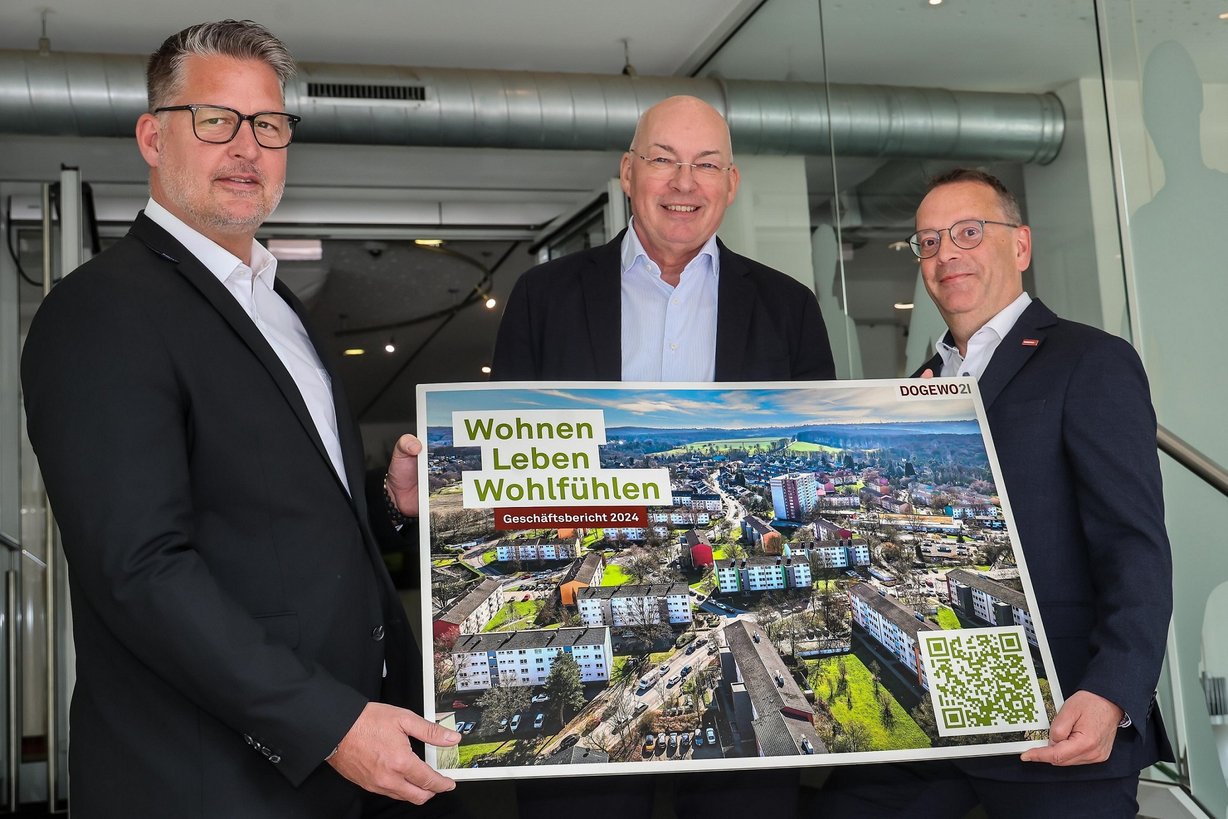 Drei Männer präsentieren im Eingangsbereich des DOGEWO21-Kundenzentrums ein großformatiges Titelbild des Geschäftsberichtes 2024.