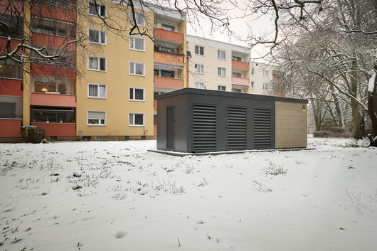 Eine Heizzentrale im Container steht vor gelb-orangene Häusern in einem schneebedeckten Garten.