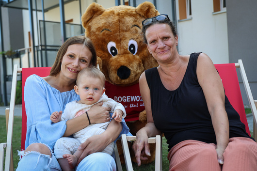 Zwei Frauen und ein Baby sitzen in roten Liegestühlen vor einem Haus, bei ihnen steht das DOGEWO21-Maskottchen Dogibär.