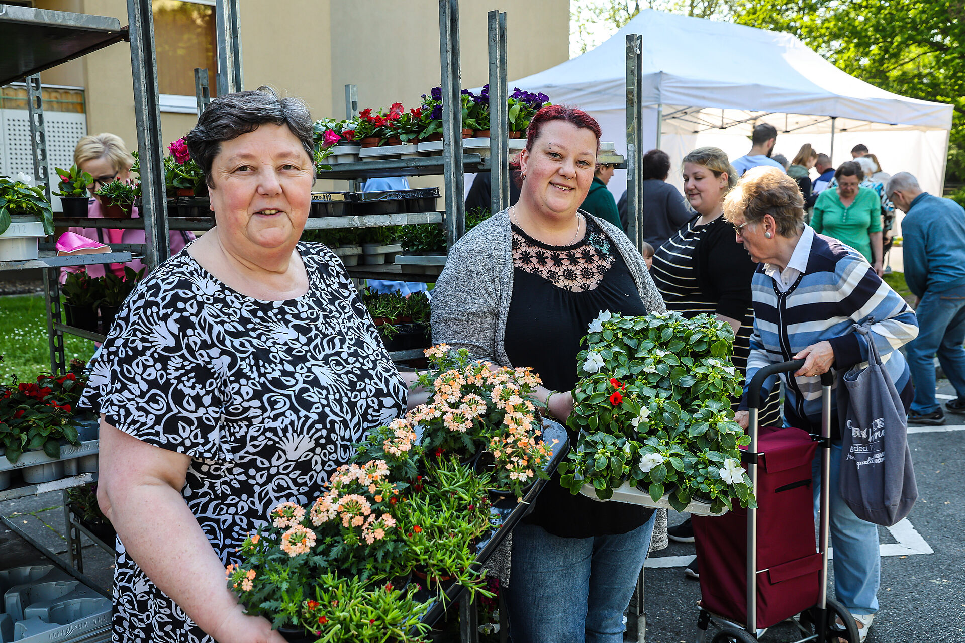 Zwei Frauen zeigen Behälter mit Balkonblumen während im Hintergrund weitere Menschen stehen