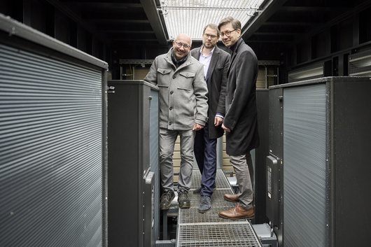 Drei Männer stehen im Innenraum eines Containers, in dem Luftwärmepumpen eingebaut sind.