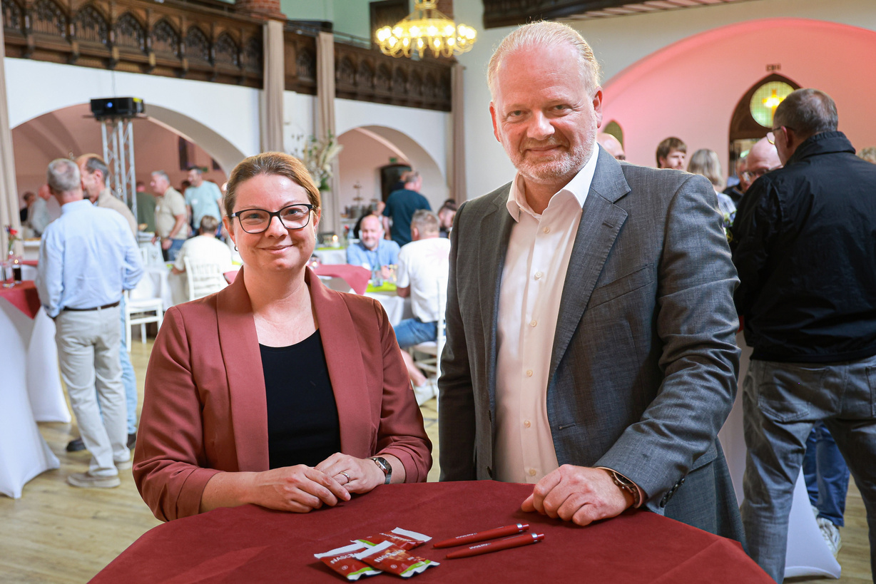 Eine Frau und ein Mann stehen lächelnd an einem Tisch, im Hintergrund sind Menschen im Gespräch miteinander.