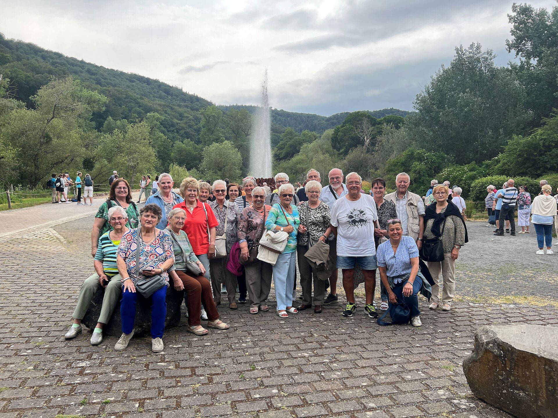 Eine Gruppe von DOGEWO21-Mieterinnen und Mietern posiert für ein Foto vor der sprühenden Wasserfontaine des Kaltwassergeysirs in Andernach