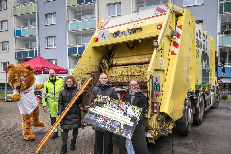 Vier Menschen und das Maskottchen Dogibär stehen vor einer gelben, mobilen Müllpresse, im Hintergrund ist ein Haus.